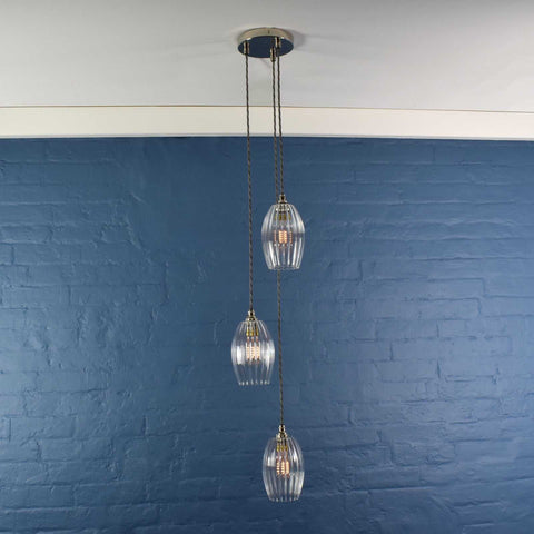Camber small ribbed glass shade cluster pendant light with three hand-blown glass shades hanging on adjustable cords against a blue brick wall background