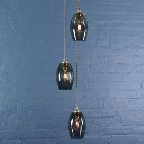Modern Camber Small Smoked Glass Shade cluster pendant light with three hand-blown smoked glass shades and adjustable flex cords against a blue brick wall background.
