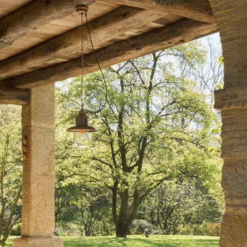 Guinguette outdoor aged brass pendant light hanging on a wooden porch with stone pillars in a garden setting