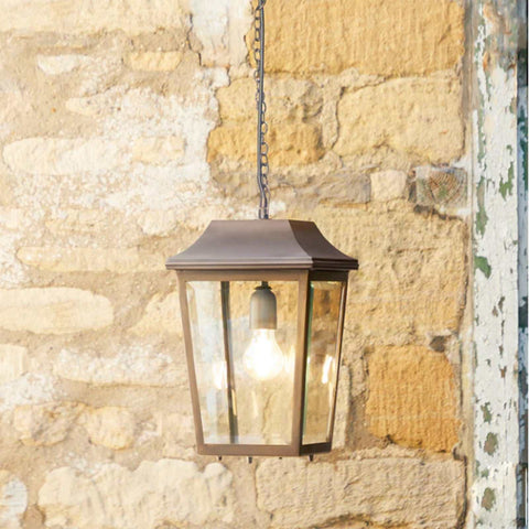 Pooky Crail Lantern outdoor ceiling light with solid brass frame and bevelled glass panels hanging on chain against stone wall