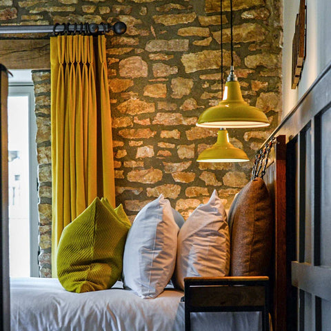 Vienna Pendant Light with mustard yellow shade hanging above a cozy seating area with green and white pillows against a rustic stone wall