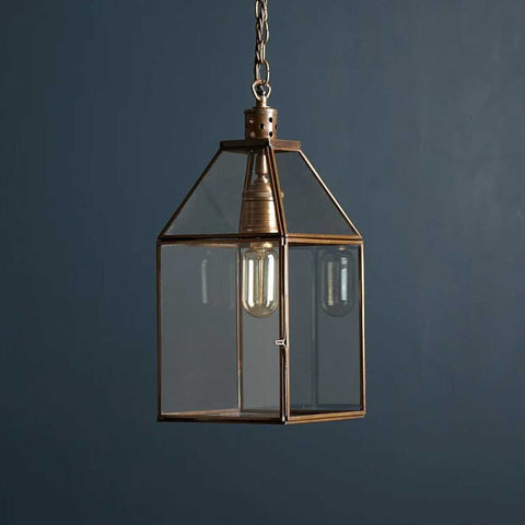 Medium Carrington Lantern Pendant Light with antique brass frame and clear glass shade hanging against a dark blue background