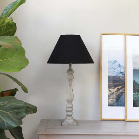 Whitwell Wooden Table Lamp with antique grey wooden base and black fabric lampshade on a table with framed wall art and green plant nearby