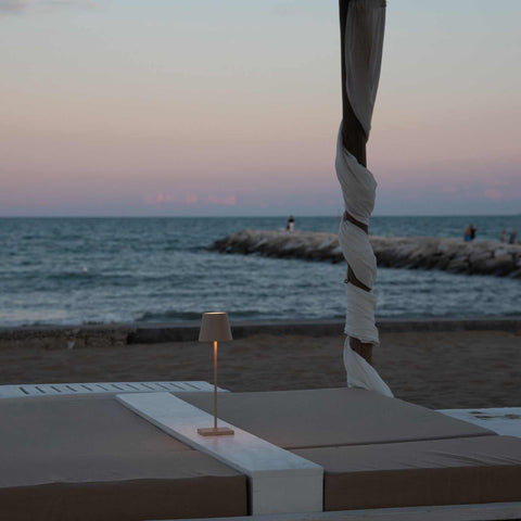 Zafferano Poldina Pro rechargeable sand table lamp glowing on an outdoor beach lounge at dusk