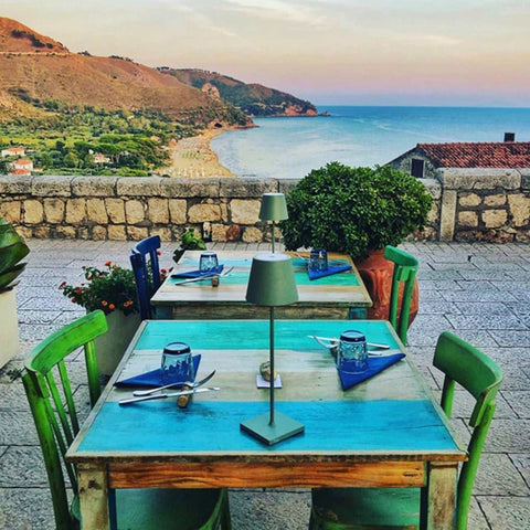 Outdoor dining table set with Zafferano Poldina Pro rechargeable sage green table lamps overlooking coastal landscape
