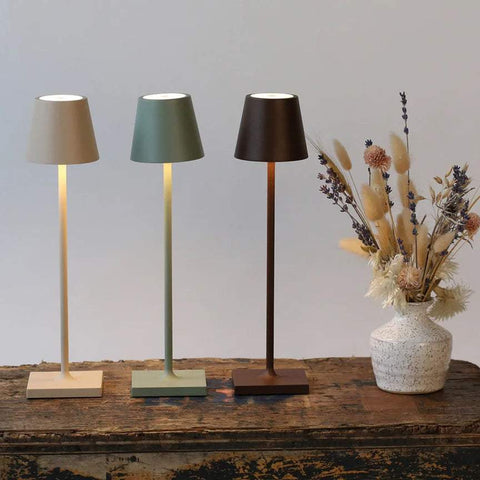 Three Zafferano Poldina Micro rechargeable LED table lamps in beige, sage green, and dark brown on a wooden surface next to a vase with dried flowers