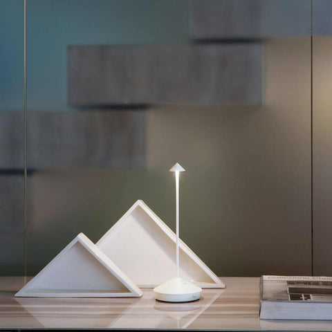 Zafferano Pina Pro rechargeable white table lamp with minimalist modern design on wooden surface next to decorative triangular objects and book