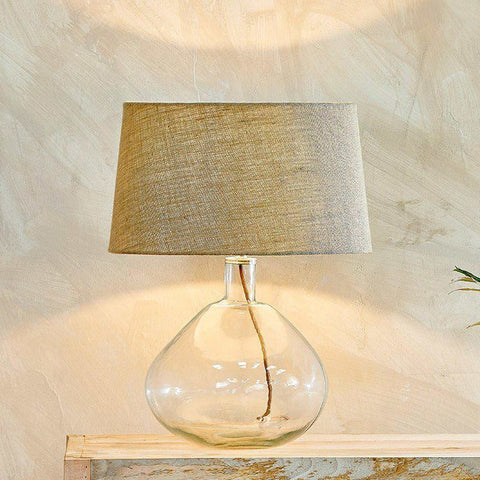 Nkuku Baba Clear Glass Table Lamp with wide rounded base and textured fabric shade on wooden surface