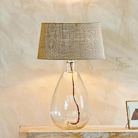 Nkuku Baba Clear Glass Table Lamp with textured fabric shade and recycled glass base on wooden surface