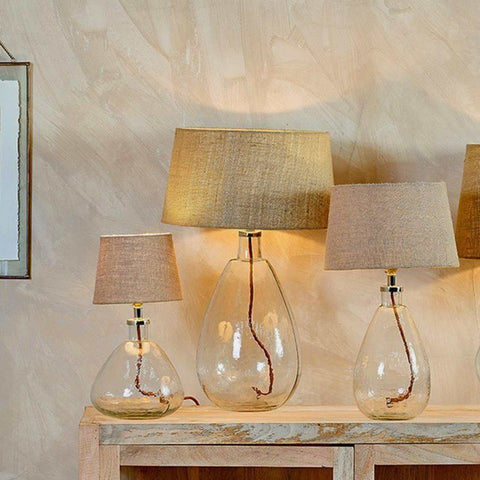 Nkuku Baba clear glass table lamp with textured fabric shade and brown braided cable on wooden surface