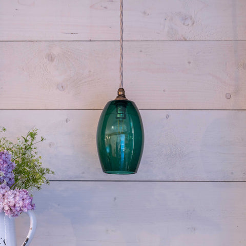 Camber Small Teal Glass pendant light hanging against a white wooden wall with a vase of purple flowers nearby