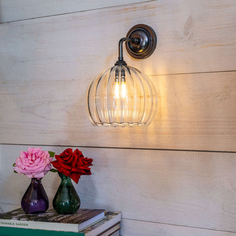 Soho Medium Ribbed Glass Shade Wall Light with bronze finish and swan neck design illuminated on wooden wall above vases and books