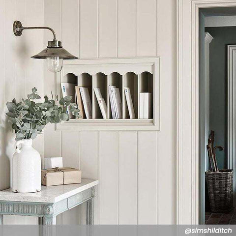 Wheston Wall Light by Chehoma in antique silver metal with industrial design mounted on white wooden wall above table with vase and books.