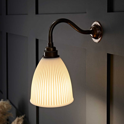 Hartlip small ribbed ceramic shade wall light with warm glow and bronze curved arm mounted on dark paneled wall
