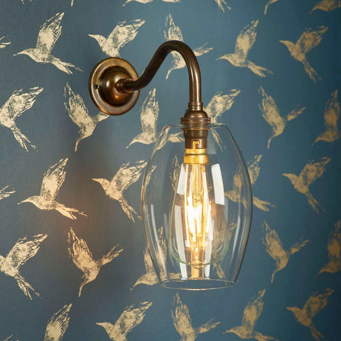 Camber Small Clear Glass Shade wall light with elongated glass shade and antique brass finish mounted on bird-patterned wallpaper background