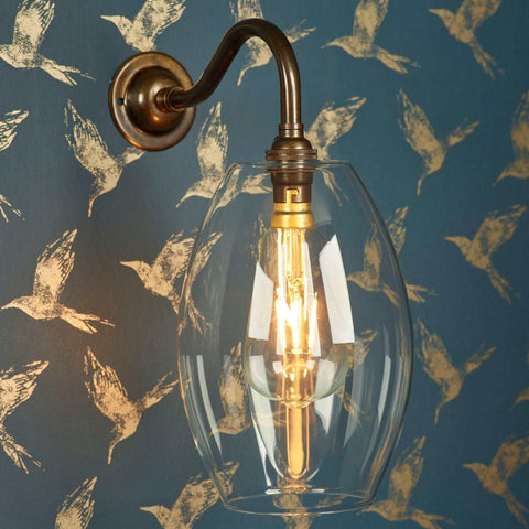 Camber Medium Clear Glass Shade wall light with vintage brass arm and clear bulb against bird-patterned blue wallpaper