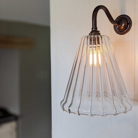 Rye Medium Ribbed Glass wall light with vintage brass arm and clear ribbed glass shade illuminating warm light on white wall