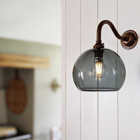 Soho Small Smoked Glass Shade wall light with vintage filament bulb and curved metal arm mounted on white wooden wall