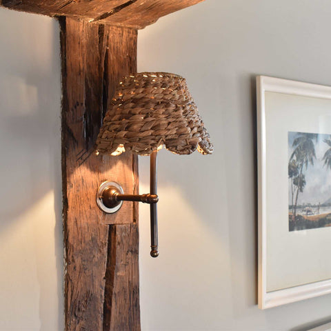 Natural seagrass shade wall light with antique brass fitting mounted on wooden beam, emitting soft warm glow in cozy room setting