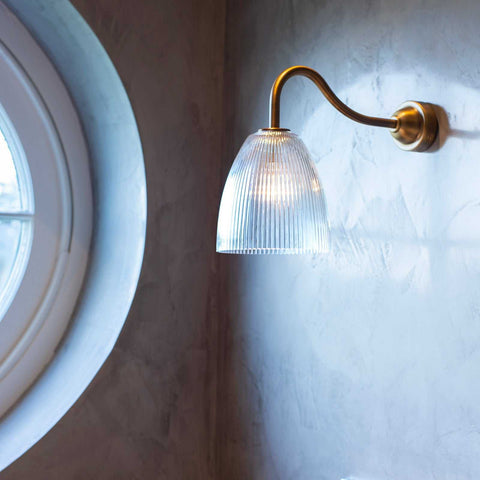 Pooky Tiny Qilin bathroom wall light with swan neck brass arm and prismatic glass shade illuminating textured wall near round window