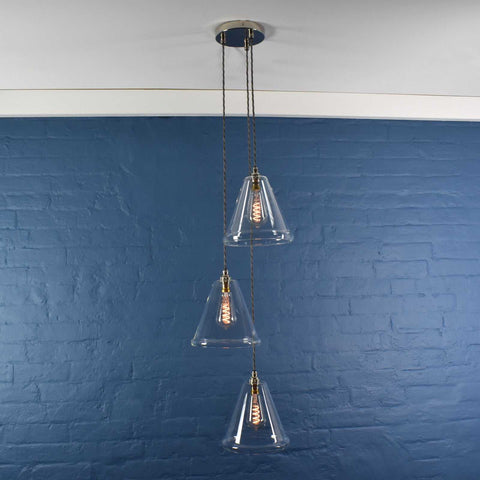 Rye medium clear glass shade cluster pendant light with three hand-blown glass shades and adjustable hanging cords against a blue brick wall background