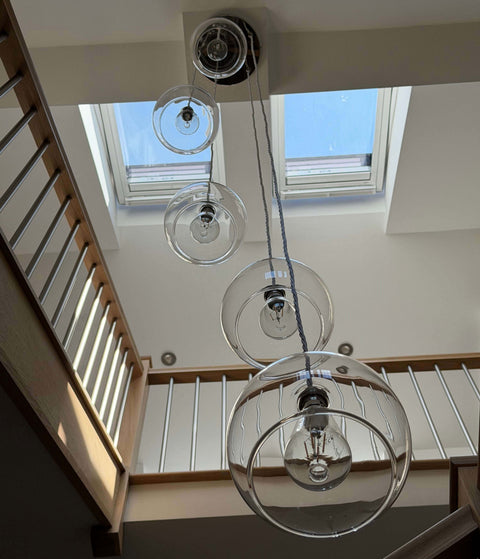 A modern multi-light pendant chandelier with four clear glass globe shades of varying sizes and black metal hardware, hanging from the ceiling of a bright home with skylights and a wooden staircase railing.