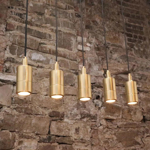 Five modern Ennis IP65 Spot Pendant Lights with sleek brass tubular design hanging from black cables against a rustic stone wall background.