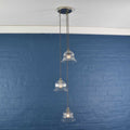 Bodium Small Ribbed Glass Shade cluster pendant light with three clear hand-blown glass shades hanging at varied lengths against a blue brick wall