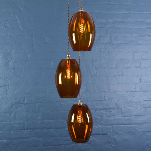 Camber large amber glass shade cluster pendant light with three oval amber glass shades hanging against a blue brick wall