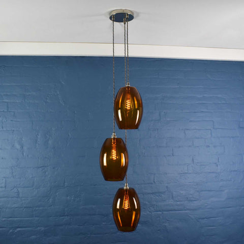 Camber large amber glass shade cluster pendant light with three handmade oval amber glass shades hanging against a blue brick wall background
