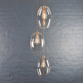 Camber large clear glass shade cluster pendant light with three handmade oval glass shades and exposed filament bulbs against a gray brick wall background