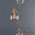 Camber medium clear glass shade cluster pendant light with three oval glass shades and vintage filament bulbs against a gray brick wall background