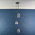 Camber small ribbed glass shade cluster pendant light with three hand-blown glass shades hanging on adjustable cords against a blue brick wall background