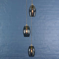 Modern Camber Small Smoked Glass Shade cluster pendant light with three hand-blown smoked glass shades and adjustable flex cords against a blue brick wall background.