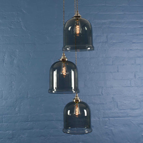 Hythe Medium Smoked Glass Shade cluster pendant light with three hand-blown smoked glass bell-shaped shades and adjustable hanging cords against a blue brick wall background