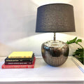 Apollo Table Lamp with antique silver hammered aluminium base and dark fabric shade on a white surface next to stacked books