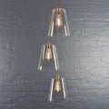 Newbury large tapered glass shade cluster pendant light with three clear hand-blown glass shades and exposed filament bulbs against a gray brick wall