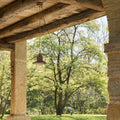 Guinguette outdoor aged brass pendant light hanging under wooden beam on patio with greenery background
