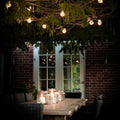 Outdoor setting with string lights hanging from branches, candles on a table, and a brick wall background.