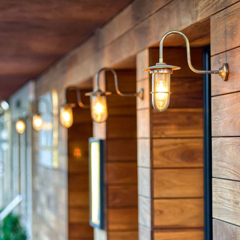 Denham Outdoor Swan Neck Wall Light in brass finish mounted on wooden exterior wall lighting walkway at dusk
