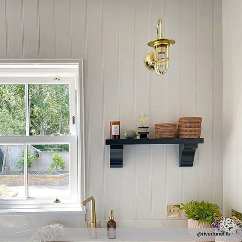 Denham Outdoor Swan Neck Wall Light in polished brass mounted on white paneled wall above black shelf in bright kitchen setting