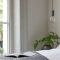 Modern Scandi Simple Bulb Pendant Light hanging beside a window in a minimalist room with a plant and an open book on a bed