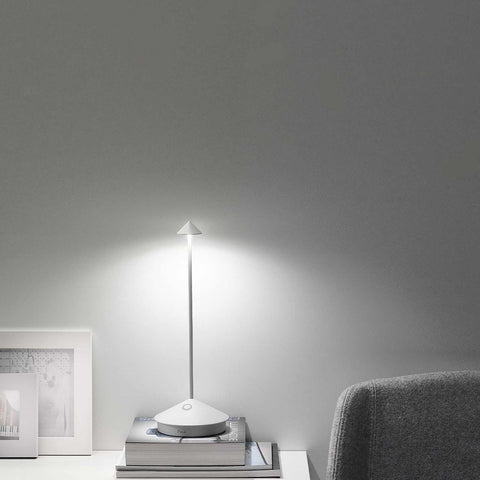 Zafferano Pina Pro rechargeable white table lamp with minimalist design on a stack of books, providing soft indoor lighting next to a gray chair and framed pictures.