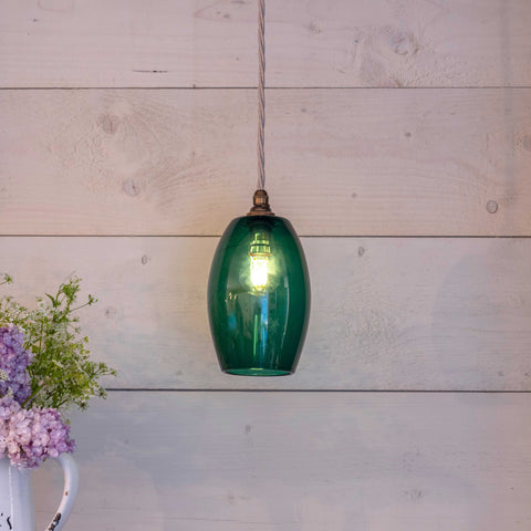 Camber Small Teal Glass pendant light hanging against a wooden wall with a visible light bulb inside and a vase of flowers nearby