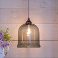 Hythe Medium Smoked Glass Shade pendant light with warm glowing bulb and adjustable hanging cord against wooden wall background