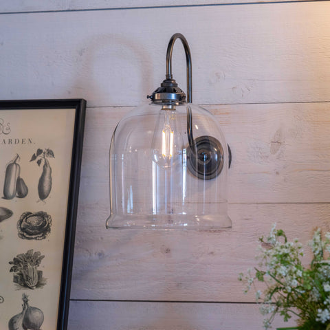Hythe Medium Clear Glass shade wall light with bell-shaped design and antique brass finish mounted on wooden wall near framed botanical print and green plant