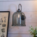 Hythe Medium Smoked Glass bell-shaped wall light with metallic finish mounted on wooden wall near framed artwork and flowers