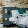 Moya Brass Wall Light with adjustable arm and cone-shaped shade mounted on a dark gray headboard beside a cozy bed with pillows and blanket in a rustic bedroom.