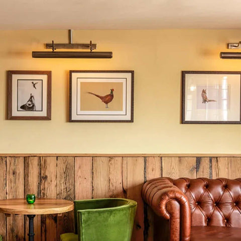 Dublin Picture Light 60.5cm solid brass wall-mounted gallery light highlighting framed bird artwork in cozy room with wooden paneling and leather sofa