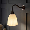 Hartlip small ribbed ceramic shade wall light with curved bronze arm and warm glow on dark paneled wall